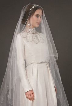 a woman wearing a white wedding gown and veil with pearls on the neckline is standing in front of a gray background