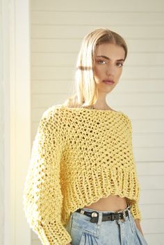 a woman wearing a yellow knitted sweater and denim shorts standing in front of a white wall