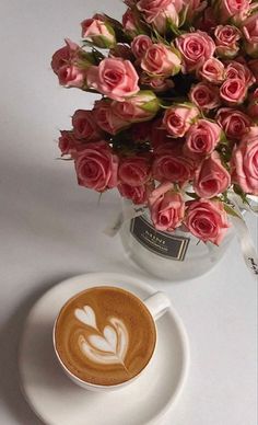 a cup of coffee next to a vase filled with pink roses on a white table