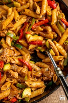 a skillet filled with pasta and vegetables on top of a wooden table next to a fork