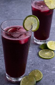 two glasses filled with red liquid and limes on the table next to each other