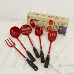 five red kitchen utensils sitting on top of a white table cloth next to a box