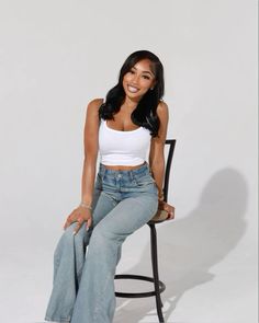 a woman sitting on top of a chair wearing high waist jeans and a white tank top