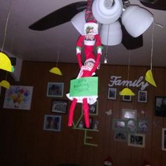 an elf hanging from a ceiling fan holding a sign