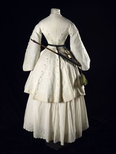 A three quarter length Marcella archery jacket; 1855. Museum of London. "A back view of a three quarter length white cotton Marcella archery jacket with pagoda sleeves, trimmed in military style with braid and covered conical buttons. Worn by Mrs. Fanny Giveen (1833-1863), the wife of Captain Xavier Giveen." 1850s Fashion, 1860 Fashion, Museum Of London, Victorian Clothing, Retro Mode, Antique Clothing, Back View