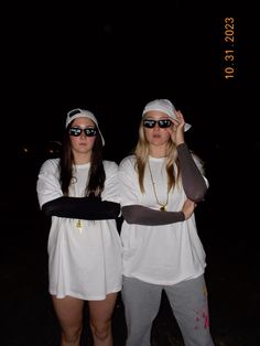 two women wearing sunglasses and white clothing standing next to each other in the dark with their arms around one another