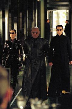 three men in black coats and sunglasses walking down a hallway