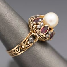 Crafted in mid-century with definite nods to Georgian design, this ring features a gorgeous glowing pearl atop bezel set rhodolite garnets separated by tiny diamond slices. The pearl is cultured and measures 7.41mm, with an ivory color and strong reflections. There are six of the rhodolite garnets- red with a pinkish hue. The garnets measure about 5 x 3mm, calculating to 0.15ct each. In between the bezels are tiny white gold floral designs holding round diamond slices. There is one on each side Georgian Design, Georgian Revival, Ring Stand, Tiny Diamond, Rhodolite Garnet, Gold Floral, Red Garnet, Ivory Color, Cultured Pearls