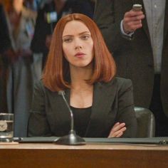 a woman with red hair sitting at a desk in front of other people and talking on the phone