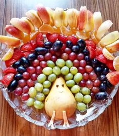 a glass plate topped with grapes, apples and oranges next to an apple shaped like a hedgehog