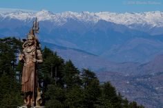 a statue in the middle of a forest with snow capped mountains behind it on a sunny day