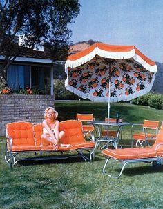 a woman sitting on top of an orange lawn chair next to an umbrella and table