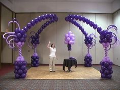 a woman is standing in front of purple balloons on the floor and an arch made out of vines