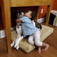 a small child is hugging a dog under a wooden table in a room with hardwood floors