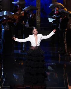 a woman in a white jacket and black skirt on stage with other people behind her