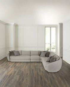 a living room with white walls and wood flooring is pictured in this image, there is a large sectional couch on the far side