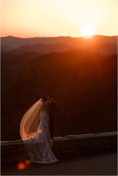 a bride and groom standing on top of a hill with the sun setting behind them