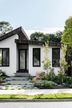 a white house with black windows and plants on the front lawn, along with rocks and grass