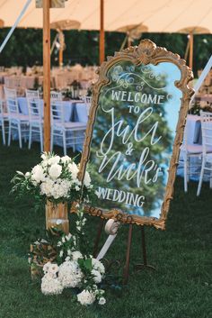 a welcome sign sitting on top of a lush green field next to a white flower filled vase