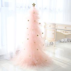 a pink christmas tree with gold stars on it in front of a white dresser and window