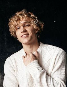 a young man with curly hair wearing a white sweater and posing for a photo in front of a black background