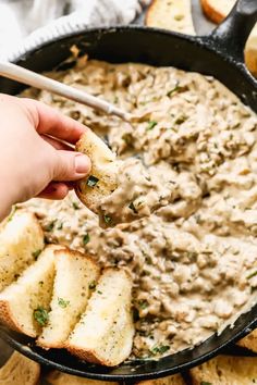 a hand holding a piece of bread over a skillet filled with dip