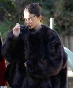 a woman in a black fur coat is looking at her cell phone while standing next to a car