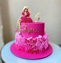 a barbie doll sitting on top of a pink cake with ruffles and gold lettering