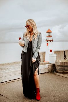 a woman in a black dress and red boots is standing near the water with a lighthouse behind her