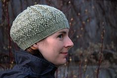 a woman wearing a green knitted hat in front of some trees and bushes with no leaves