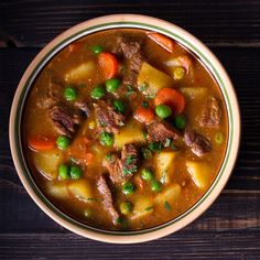 a bowl of beef stew with carrots, peas and potatoes on a wooden table