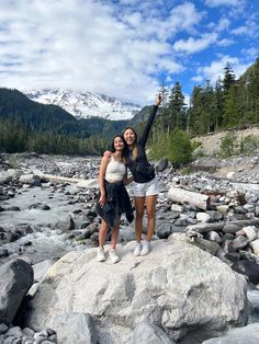 two women standing on rocks in the middle of a river with mountains in the background