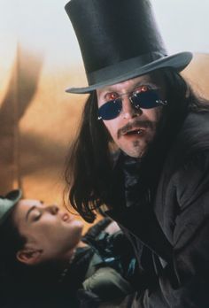 a man with long hair wearing a top hat and sunglasses next to a woman in a trench coat