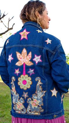 a woman wearing a blue jean jacket with stars and flowers on the back, standing in front of a tree