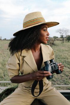 a woman wearing a hat and holding a camera in front of a field with trees