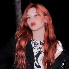 a woman with long red hair wearing a black jacket and white shirt standing in front of a tree