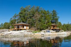 two wooden cabins sit on the shore of a body of water with trees in the background