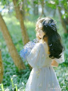 a woman in a white dress is holding blue flowers and looking at the ground with trees behind her