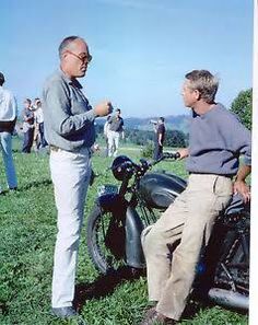 two men standing next to each other in the grass with motorcycles and people behind them