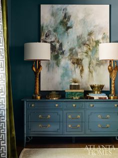 two lamps sitting on top of a blue dresser in front of a painting with gold accents