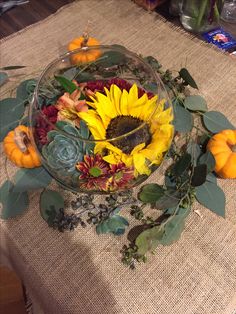 a glass bowl filled with sunflowers and greenery on top of a table