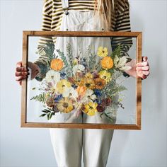 a woman holding up a framed painting with flowers in it's center and greenery on the bottom