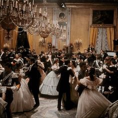 a group of people dressed in formal dress dancing together at a ball with chandeliers hanging from the ceiling