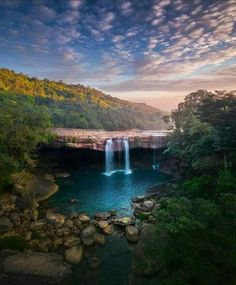 a large waterfall in the middle of a lake surrounded by trees and rocks under a cloudy blue sky