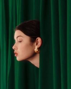 a woman with her eyes closed looking out from behind a green curtain, wearing gold earrings