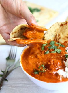 a hand holding a tortilla chip over a bowl of red curry and rice