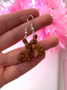 a hand holding two small brown dogs on it's earring hooks, with pink flowers in the background