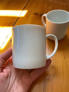 a hand holding a white coffee cup on top of a wooden table next to another cup