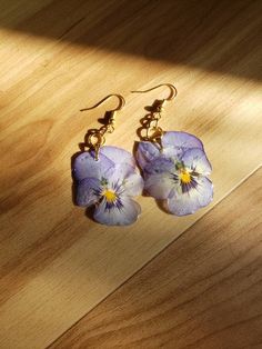 two purple pansies are hanging from gold earwires on a wooden surface in the sunlight