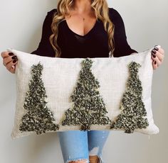 a woman holding up a pillow with christmas trees on the front and sides, while wearing ripped jeans